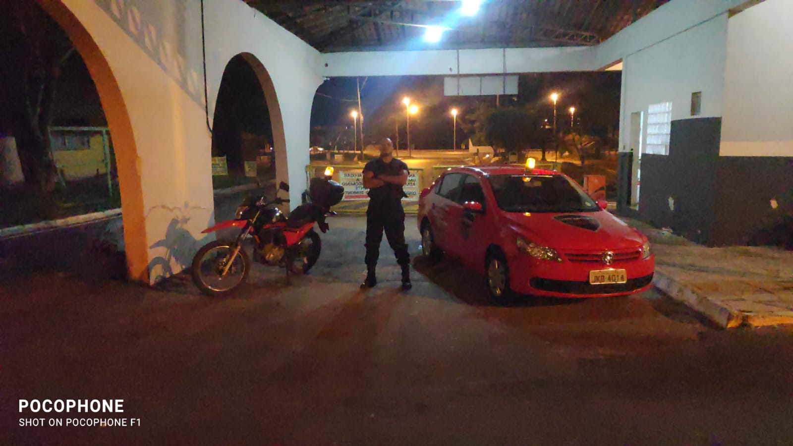 Segurança em frente à entrada durante ronda noturna com moto e carro visíveis
