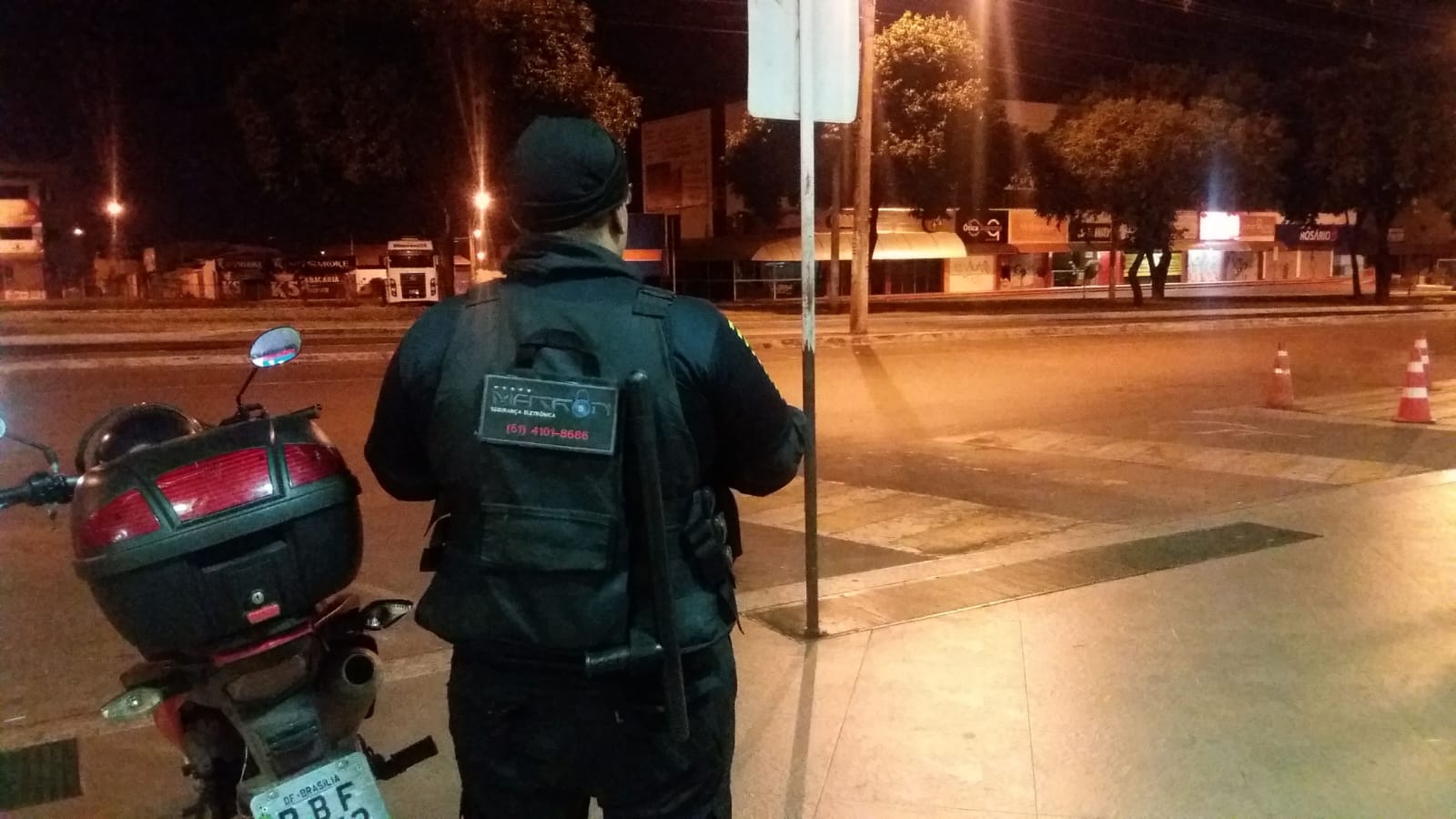 Vigilante da Matron em ronda noturna com colete tático ao lado de moto de apoio